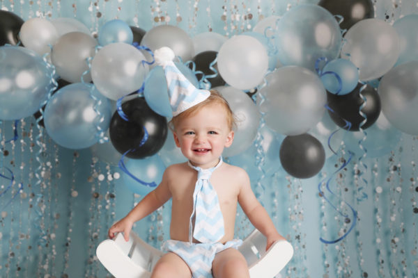 first birthday cakesmash baby boy smiling in party hat with blue balloon backdrop