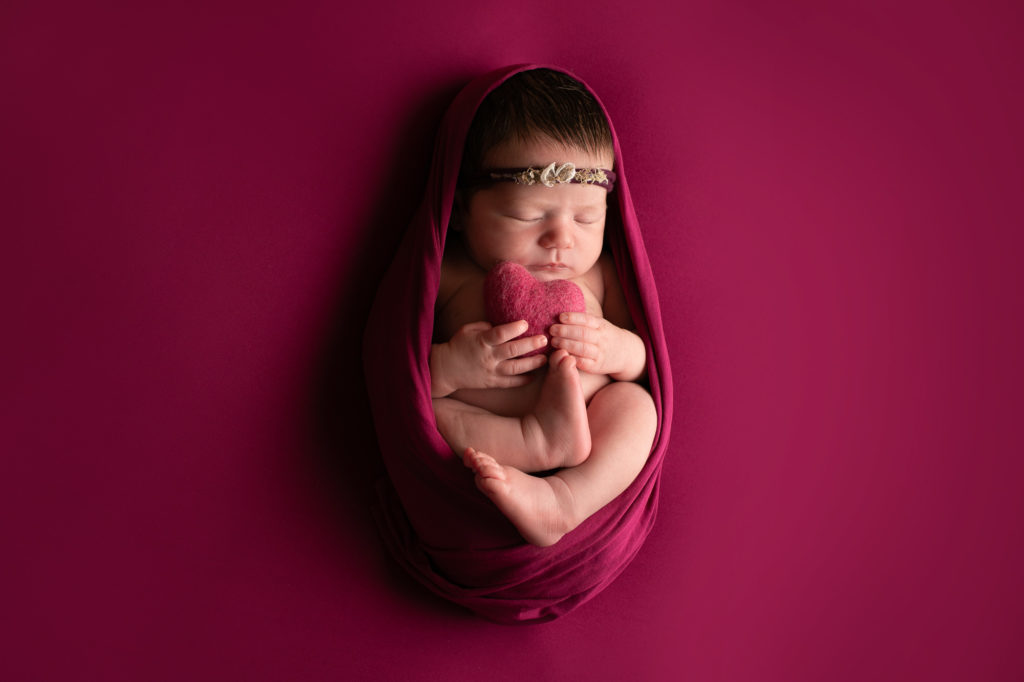 newborn baby girl portrait in deep pink rap holding small pink heart